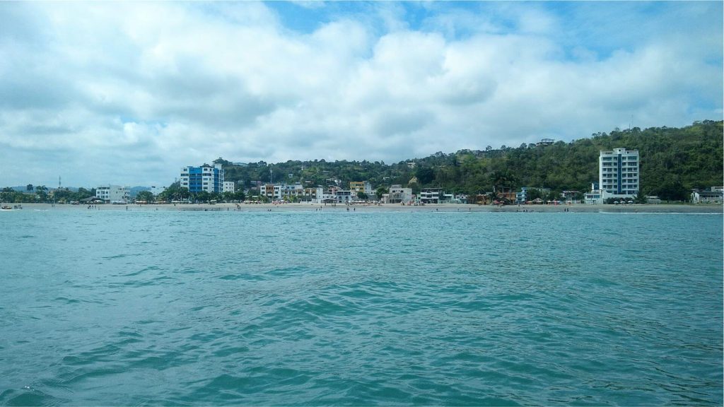 Un mar turquesa y tranquilo, con suaves olas en primer plano. La costa lejana de Atacames presenta edificios, árboles frondosos y un cielo nublado, evocando una atmósfera costera serena.