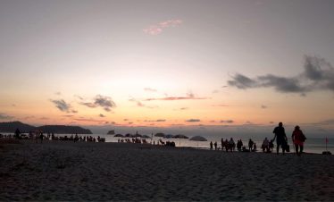 Siluetas de personas pasean por la playa de Atacames al atardecer, con suaves tonos rosas y naranjas en el cielo. Sombrillas y gente salpican la orilla, creando una escena tranquila.