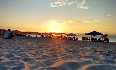Atardecer en una playa de arena con siluetas dispersas de personas bajo sombrillas. El cielo luce una cálida mezcla de naranja y rosa, creando una atmósfera serena y relajada.
