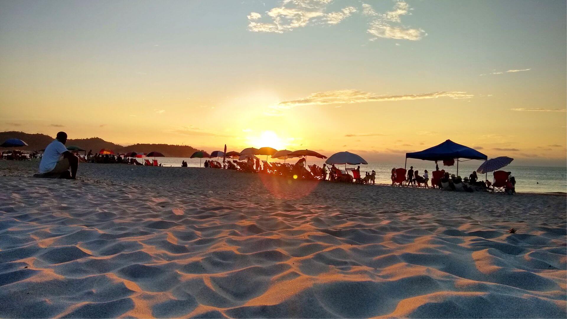 Atardecer en una playa de arena con siluetas dispersas de personas bajo sombrillas. El cielo luce una cálida mezcla de naranja y rosa, creando una atmósfera serena y relajada.