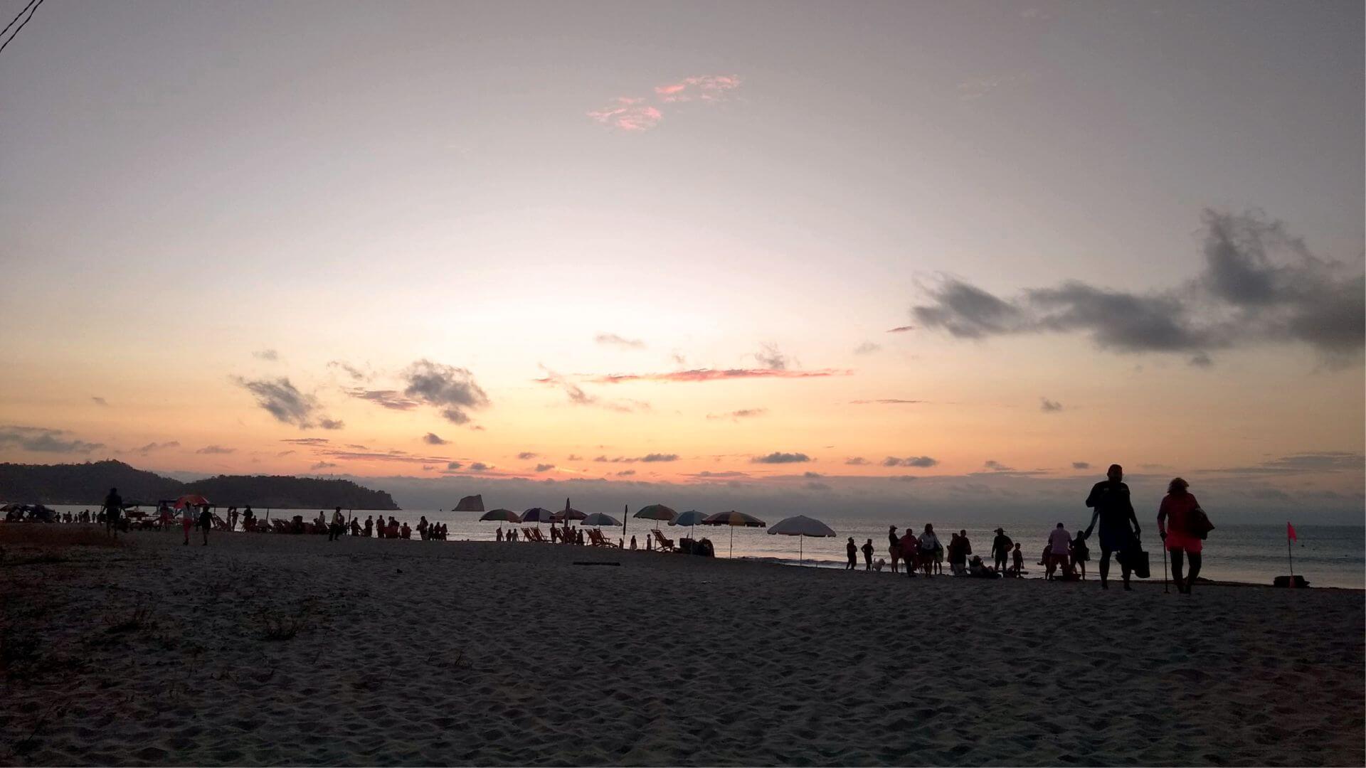 Siluetas de personas pasean por la playa de Atacames al atardecer, con suaves tonos rosas y naranjas en el cielo. Sombrillas y gente salpican la orilla, creando una escena tranquila.