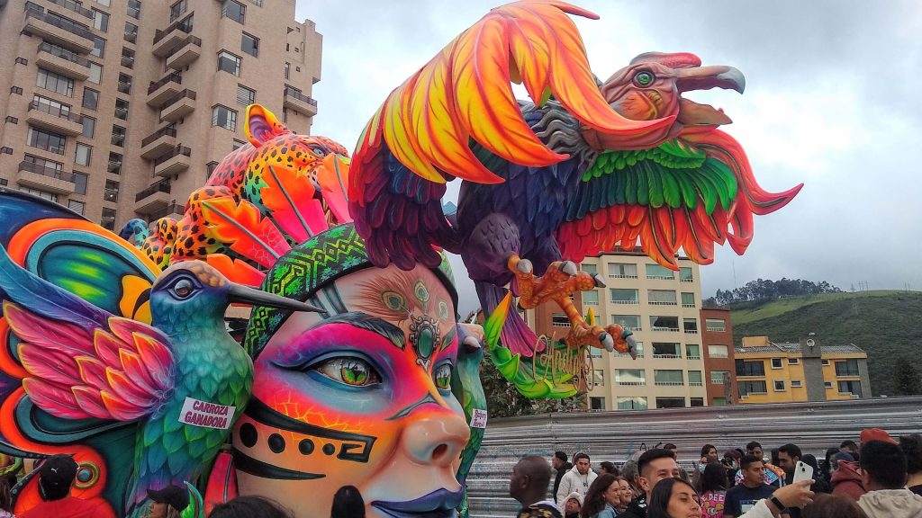 Carroza vibrante con una máscara colorida y un condor de tamaño, rodeada de multitud. Tonos brillantes y ambiente festivo, con edificios de la ciudad al fondo.