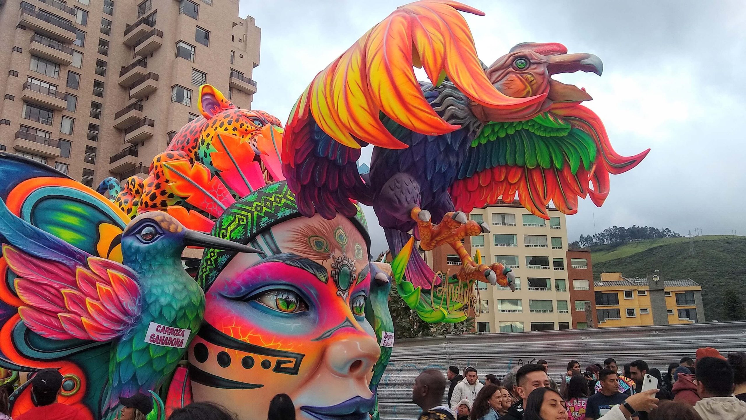 Carroza vibrante con una máscara colorida y un condor de tamaño, rodeada de multitud. Tonos brillantes y ambiente festivo, con edificios de la ciudad al fondo.