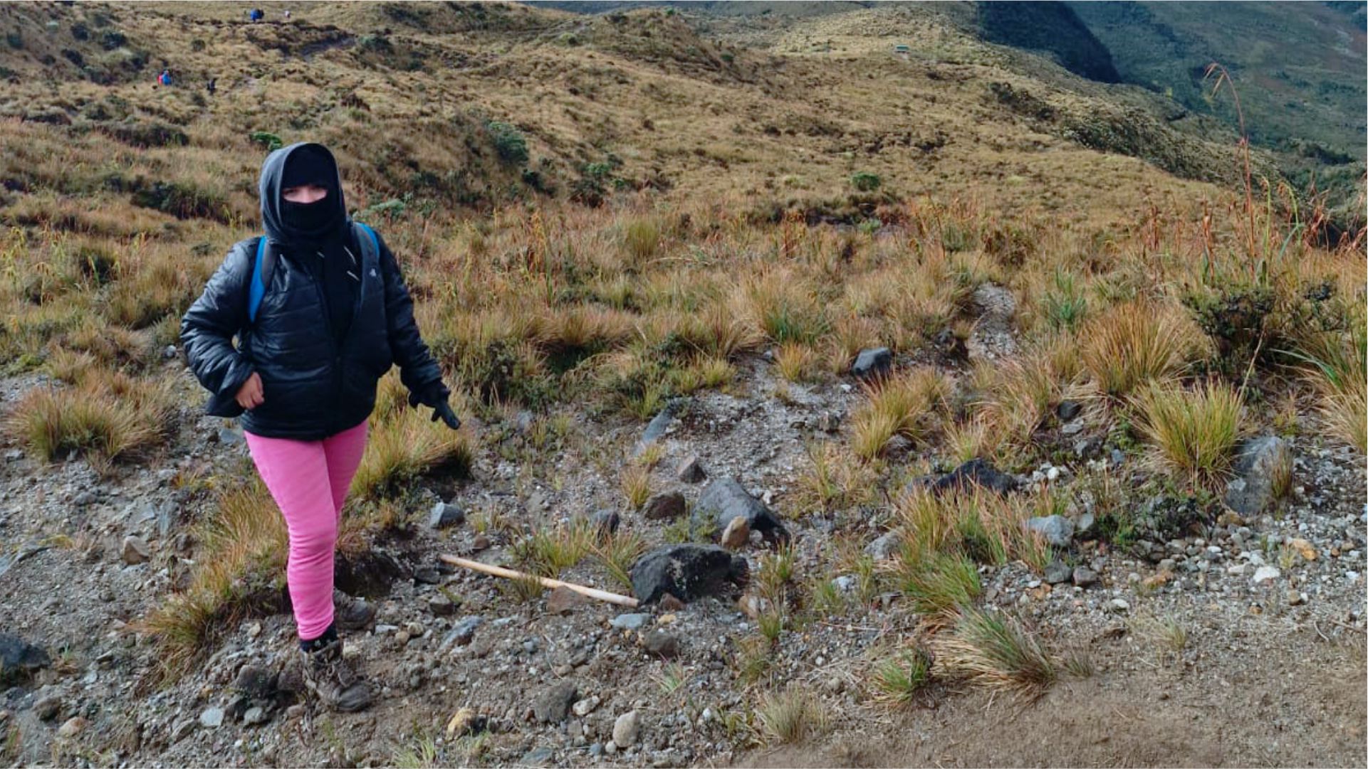 Una persona con chaqueta negra, pantalones rosas y mascarilla se encuentra de pie en un sendero rocoso y cubierto de hierba en una ladera, transmitiendo una sensación de aventura en un paisaje remoto.