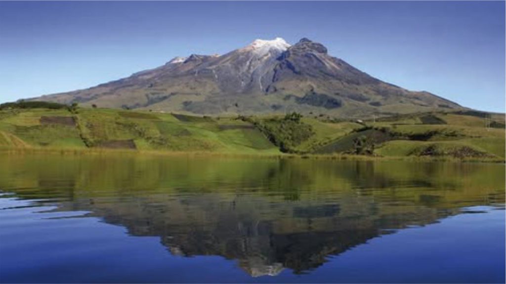 Un paisaje sereno con el volcán Cumbal con la cima nevada bajo un cielo azul despejado, reflejada a la perfección en un lago tranquilo, rodeado de exuberantes campos verdes.