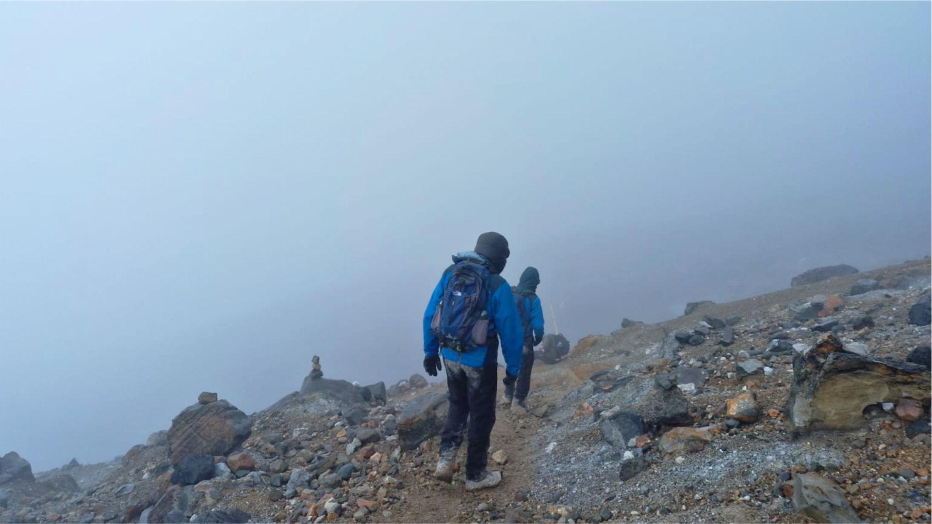 Dos excursionistas con chaquetas azules descienden por un sendero de montaña rocoso y envuelto en niebla. La atmósfera brumosa transmite una sensación de aventura y aislamiento.