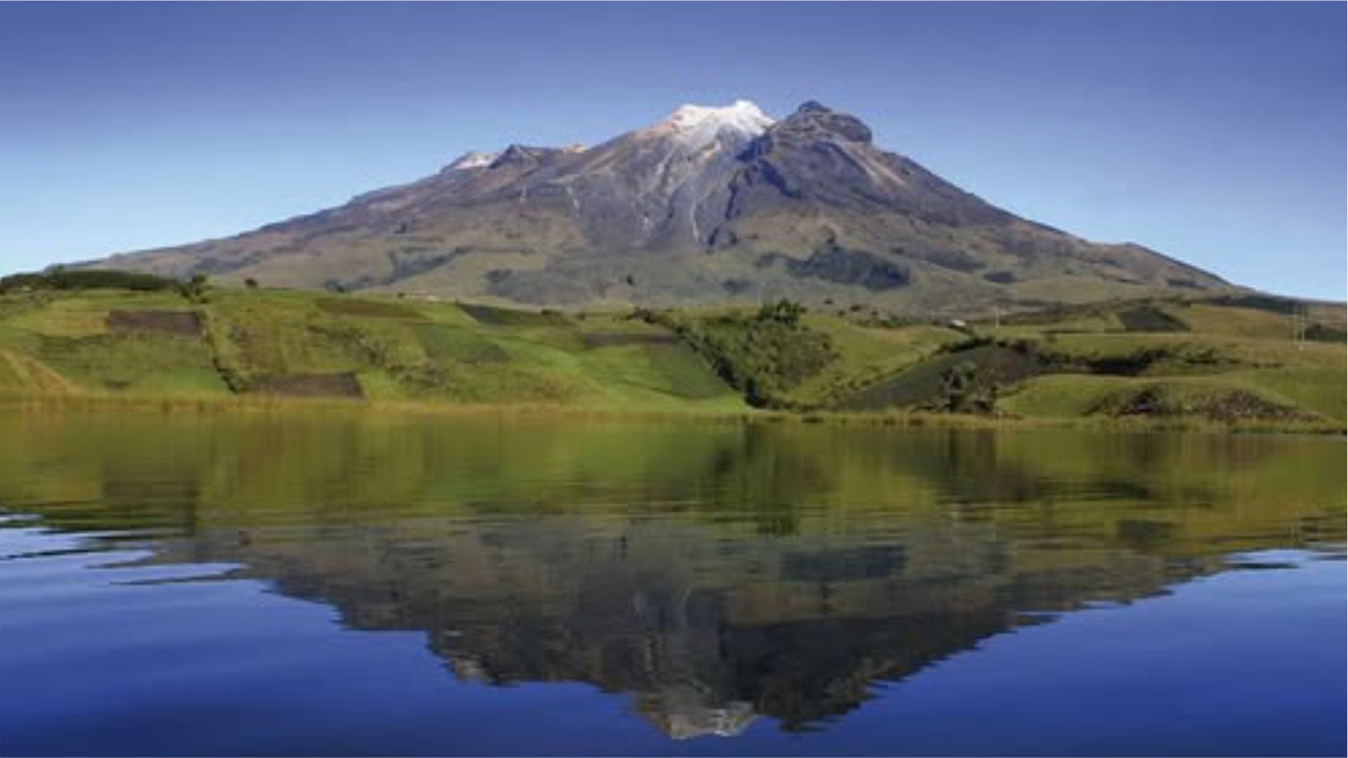 Un paisaje sereno con el volcán Cumbal con la cima nevada bajo un cielo azul despejado, reflejada a la perfección en un lago tranquilo, rodeado de exuberantes campos verdes.