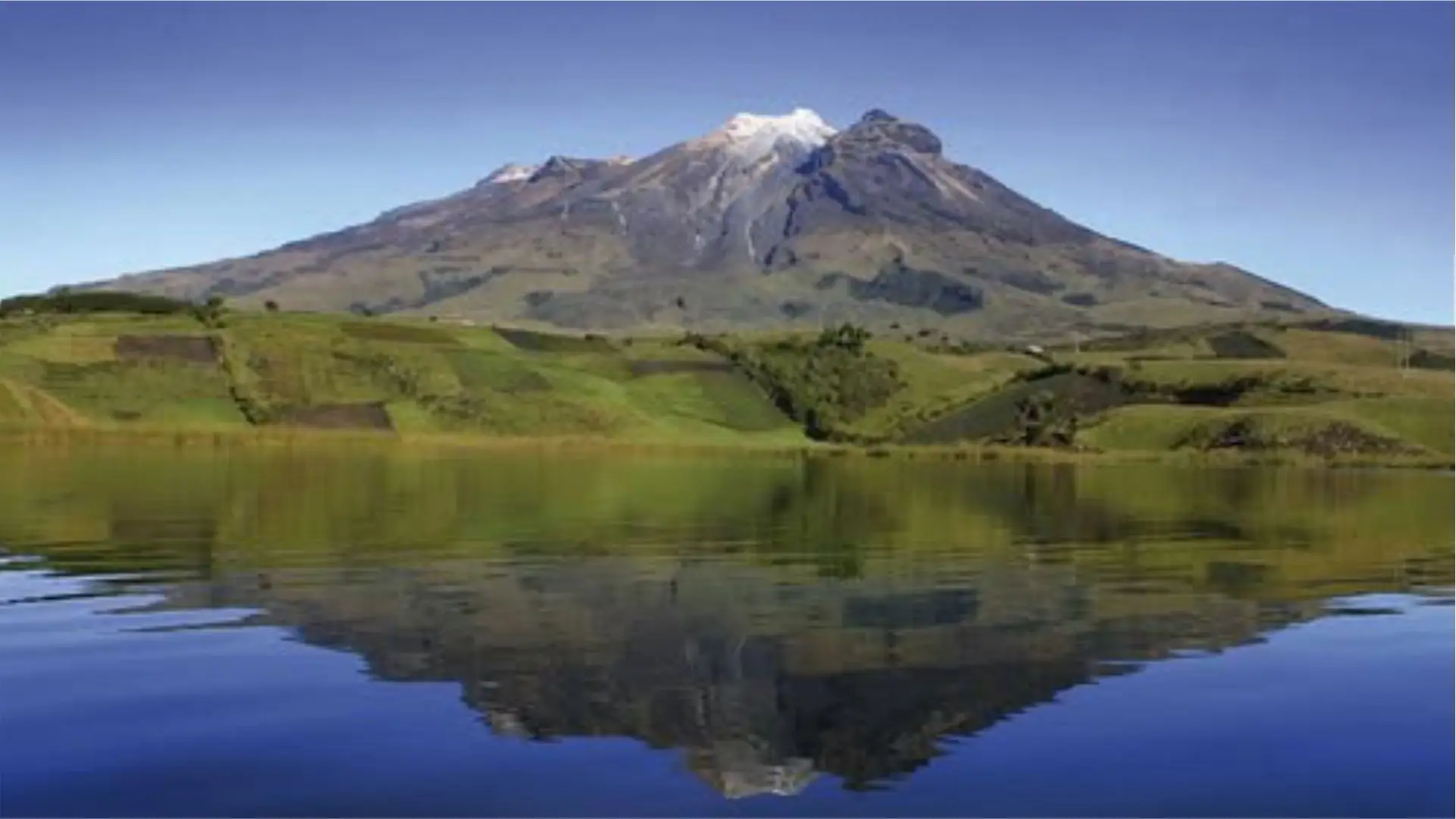 Un paisaje sereno con el volcán Cumbal con la cima nevada bajo un cielo azul despejado, reflejada a la perfección en un lago tranquilo, rodeado de exuberantes campos verdes.