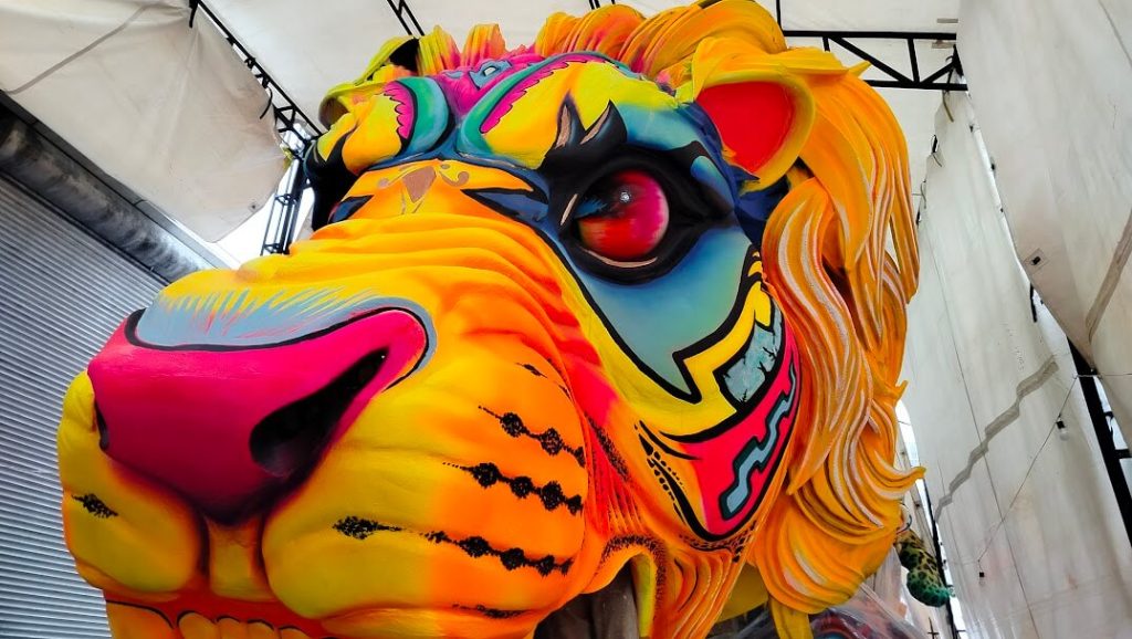 Caroza del carnaval de negros y blancos en contrucción. Máscara de león vibrante con colores neón y estampados llamativos, en intensos tonos rosa, azul y naranja. Su expresión dinámica transmite energía y entusiasmo.