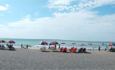 Una amplia escena playera de Atacames con coloridas sombrillas y sillas alineadas cerca del mar. La gente disfruta de diversas actividades bajo un cielo azul parcialmente nublado, creando un ambiente animado y relajado.