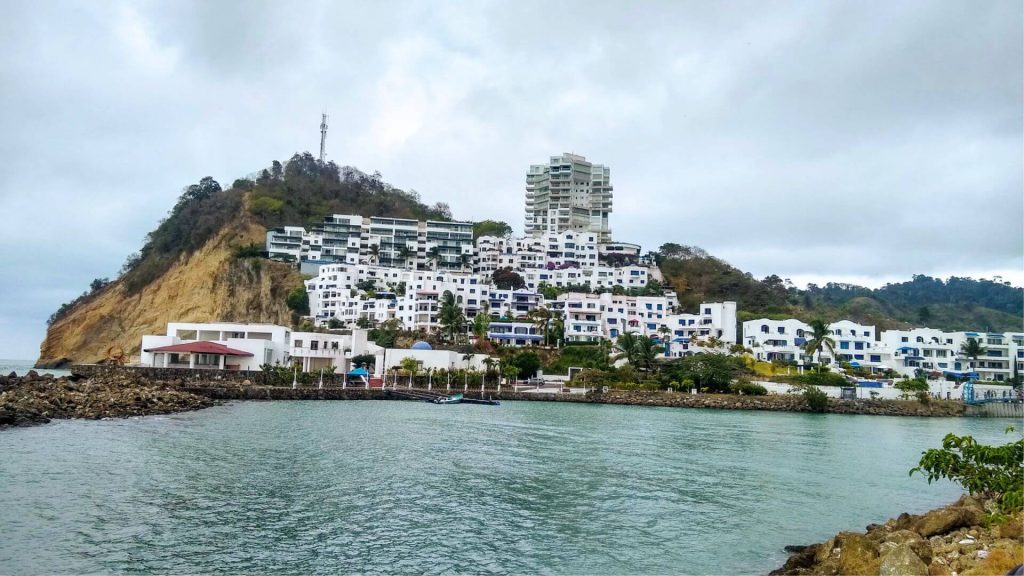 Edificios blancos con detalles azules se agrupan en una ladera verde junto a una costa rocosa, bajo un cielo nublado, creando una escena costera apacible.