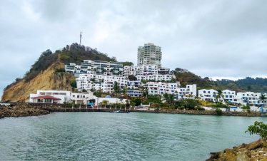 Edificios blancos con detalles azules se agrupan en una ladera verde junto a una costa rocosa, bajo un cielo nublado, creando una escena costera apacible.