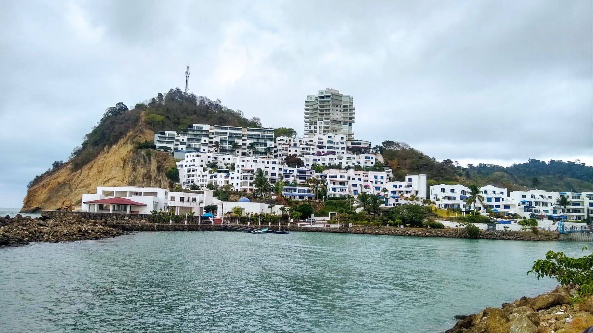 Edificios blancos con detalles azules se agrupan en una ladera verde junto a una costa rocosa, bajo un cielo nublado, creando una escena costera apacible.