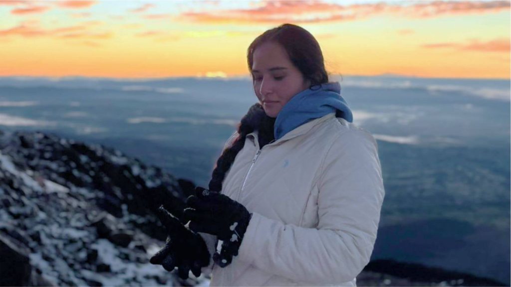 Una persona con bata y guantes blancos se encuentra de pie en una montaña cubierta de nieve al amanecer, con una vista panorámica y un sereno cielo naranja de fondo.
