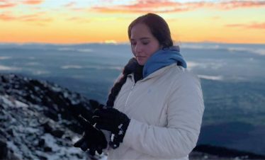 Una persona con bata y guantes blancos se encuentra de pie en una montaña cubierta de nieve al amanecer, con una vista panorámica y un sereno cielo naranja de fondo.