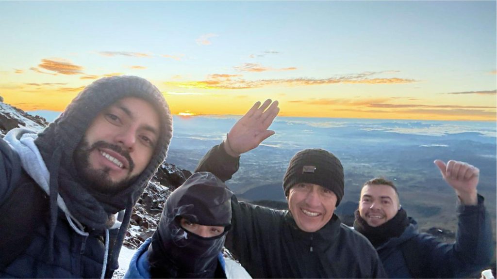 Un grupo de cuatro personas abrigadas con ropa de invierno sonríen a la cámara en una montaña nevada al amanecer, con un impresionante paisaje de fondo.