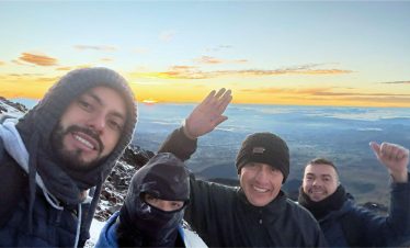 Un grupo de cuatro personas abrigadas con ropa de invierno sonríen a la cámara en una montaña nevada al amanecer, con un impresionante paisaje de fondo.