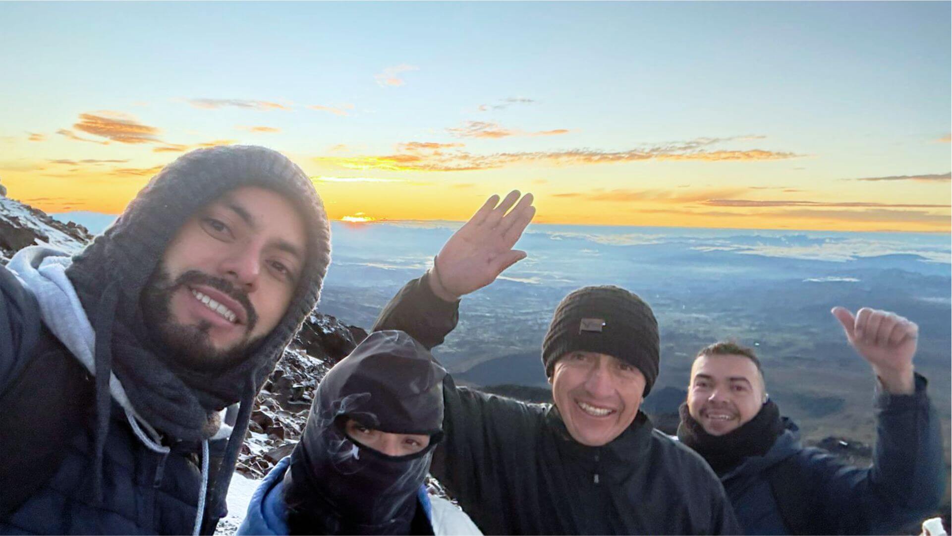 Un grupo de cuatro personas abrigadas con ropa de invierno sonríen a la cámara en una montaña nevada al amanecer, con un impresionante paisaje de fondo.