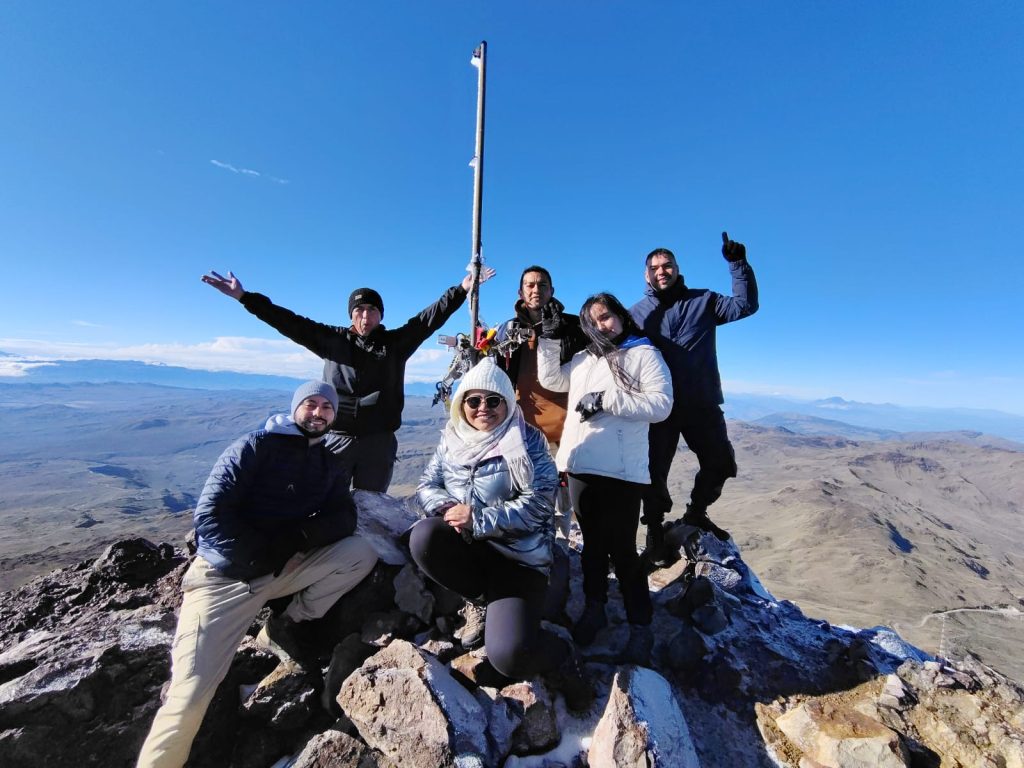 Un grupo de seis personas con ropa de invierno celebra en la cima de una montaña bajo un cielo azul despejado. Sonríen, posan con confianza y derrochan alegría.