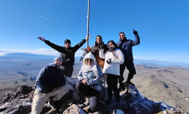 Un grupo de seis personas con ropa de invierno celebra en la cima de una montaña bajo un cielo azul despejado. Sonríen, posan con confianza y derrochan alegría.