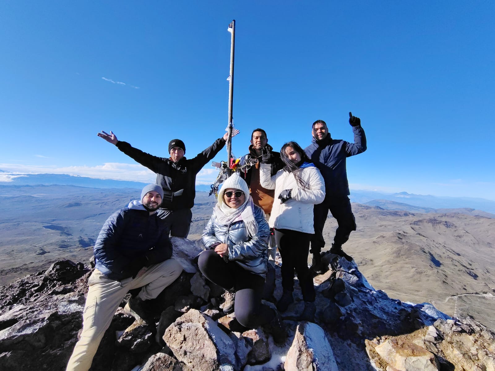 Un grupo de seis personas con ropa de invierno celebra en la cima de una montaña bajo un cielo azul despejado. Sonríen, posan con confianza y derrochan alegría.