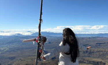 Una mujer con chaqueta blanca está de pie con los brazos extendidos en la cima del Volcán del Chiles junto a una cruz decorada. Un vasto paisaje bajo un cielo azul despejado.