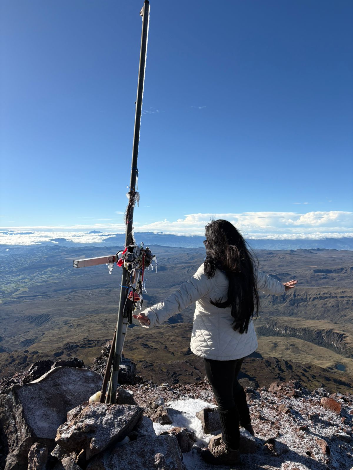 Una mujer con chaqueta blanca está de pie con los brazos extendidos en la cima del Volcán del Chiles junto a una cruz decorada. Un vasto paisaje bajo un cielo azul despejado.