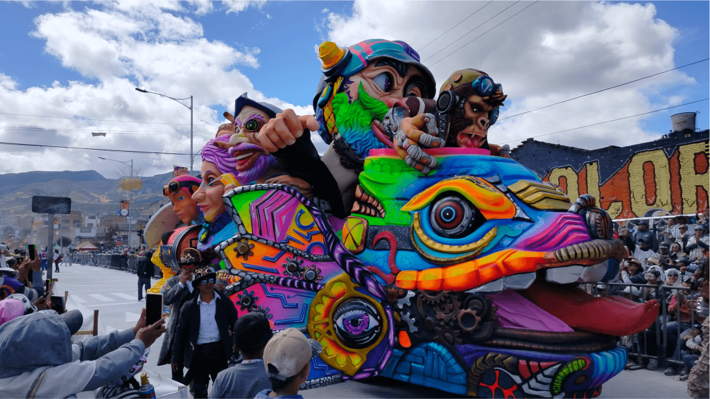 Carroza colorida con personajes caricaturescos y vibrantes que recuerdan a un camaleón y a ciclistas. La multitud se reúne bajo un cielo brillante, aunque parcialmente nublado. Ambiente festivo.