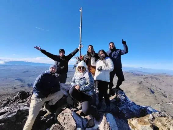 Un grupo de seis personas con ropa de invierno celebra en la cima de una montaña bajo un cielo azul despejado. Sonríen, posan con confianza y derrochan alegría.