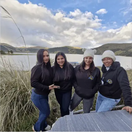 posando juntas al aire libre. Equipo de La Juana Travel, detrás de ellas, se puede apreciar un lago yLa Laguna de la Cocha; colinas verdes bajo un cielo con nubes dispersas. 
